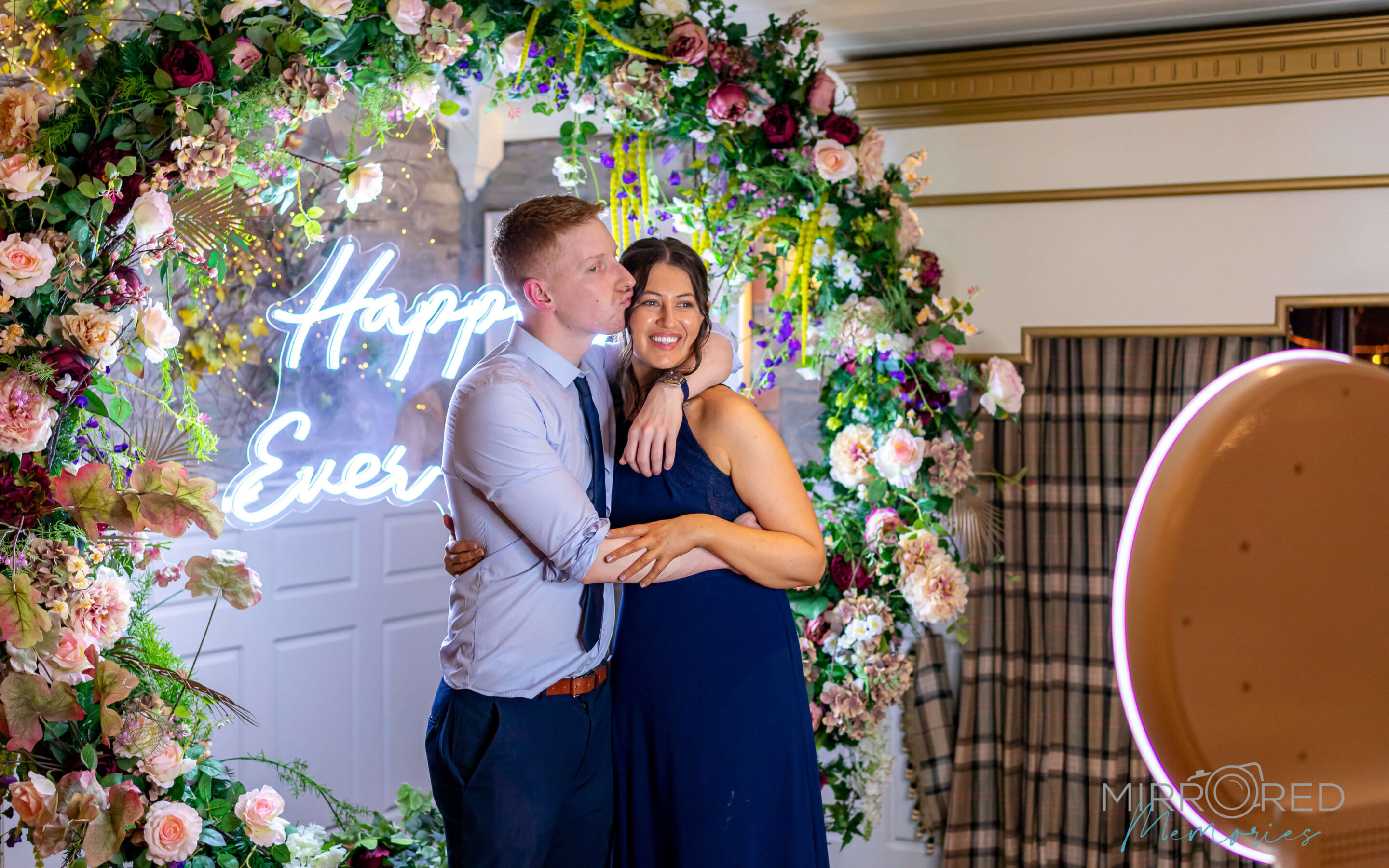 Newlyweds embracing under a floral arch with a neon Happy Ever sign next to the San Diego selfie booth